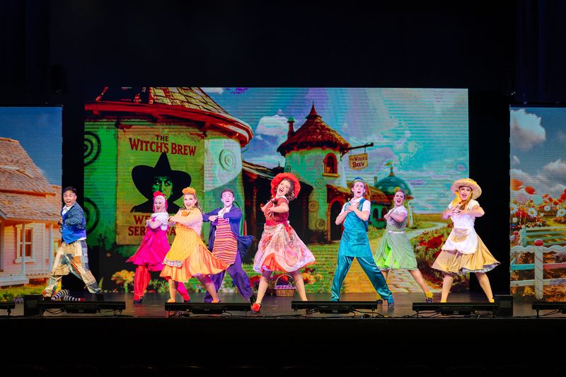 The ensemble dancing in Munchkinland in front of a colourful projected backdrop, The Wizard of Oz pantomime by Zealous Productions, December 2025